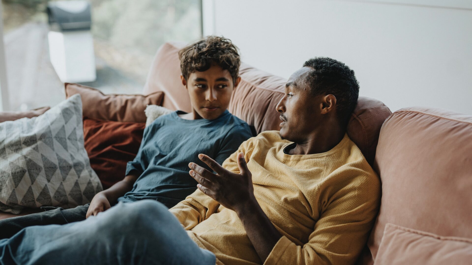 Father talking with son while sitting on sofa in living room at home - stock photo